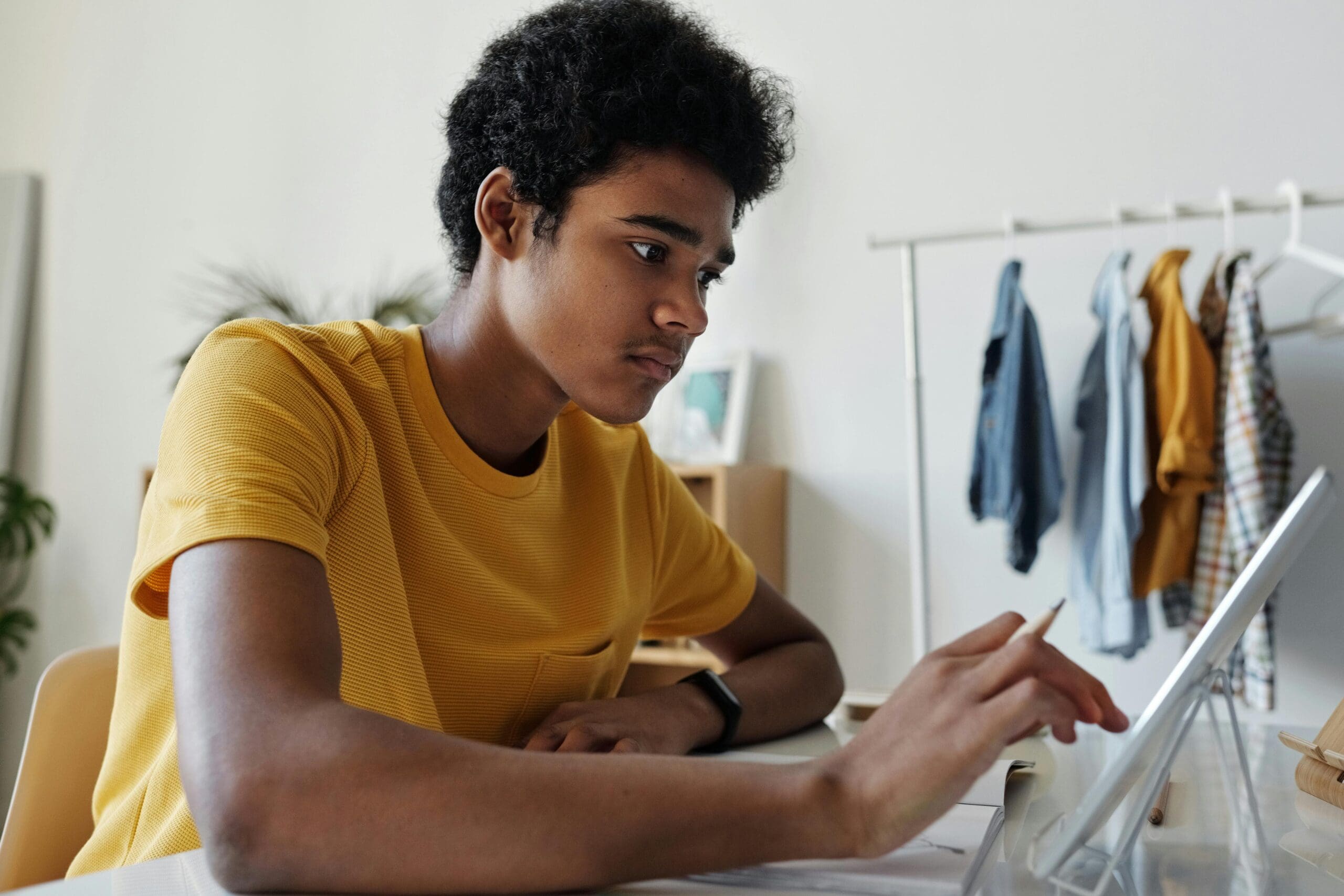 A person in a yellow shirt uses a stylus on a tablet at a desk. Clothing hangs on a rack in the background.