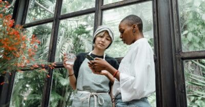 Two people stand near a large window with plants outside. One person holds a glass of water, and the other looks at a smartphone.