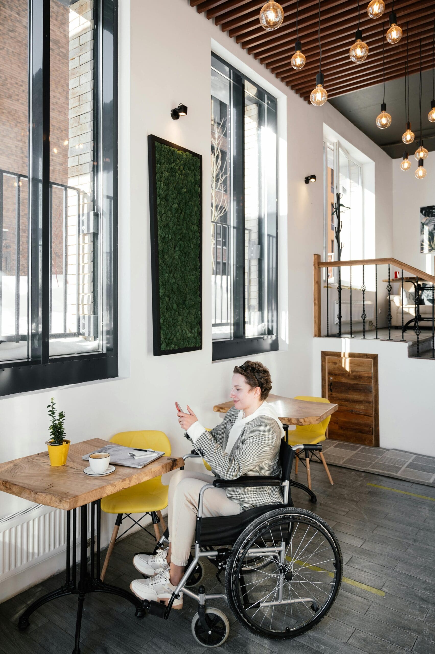 A person in a wheelchair sits at a table in a bright cafe, holding a smartphone. The table has a coffee cup, book, and plant. Large windows and hanging lights are in the background.