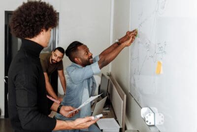Three people work together at a whiteboard, discussing and pointing out diagrams, in an office setting.