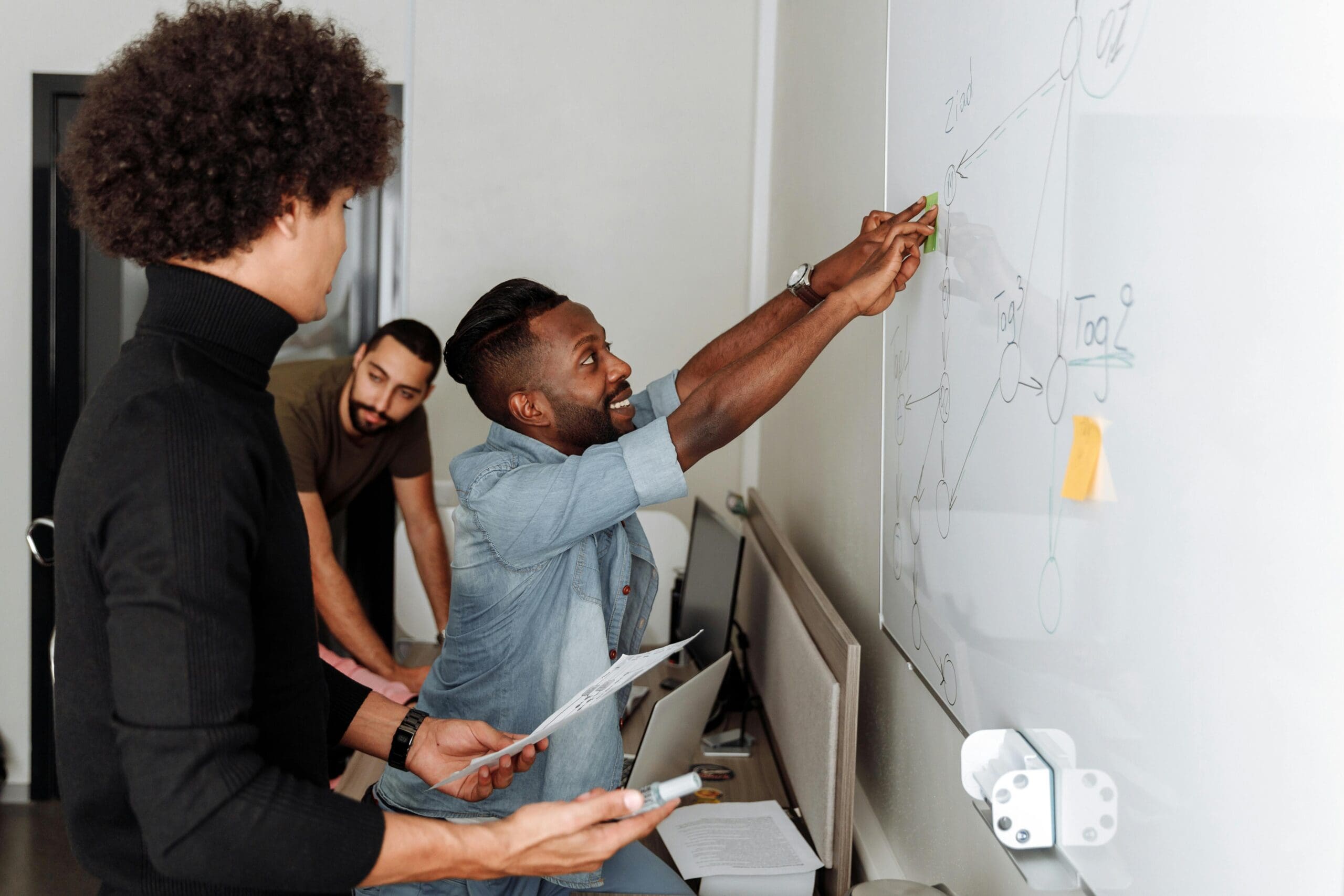 Three people work together at a whiteboard, discussing and pointing out diagrams, in an office setting.