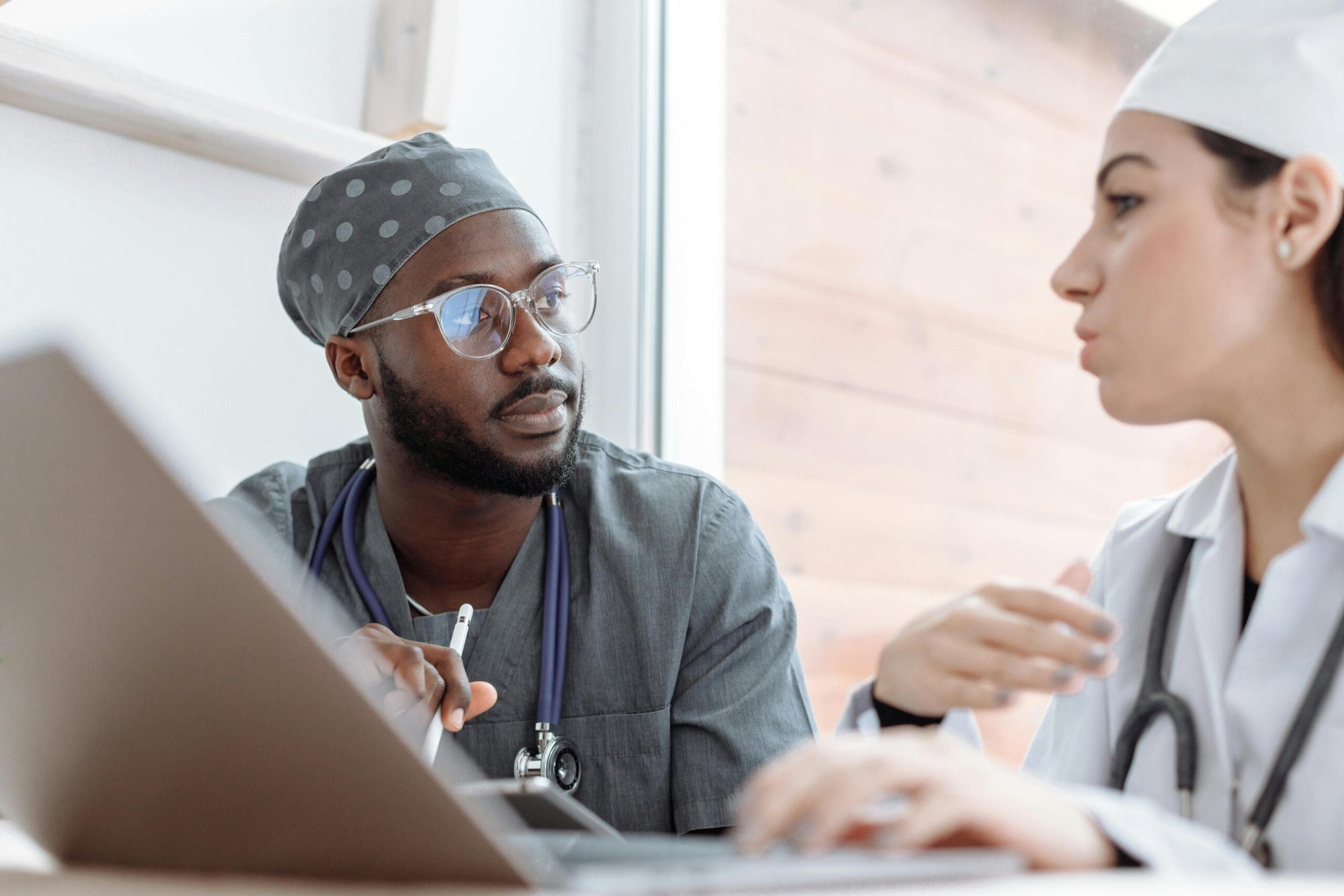 Two medical professionals in discussion, one with a laptop. Both are wearing scrubs and stethoscopes.