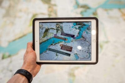 A person holds a tablet displaying a detailed map with various color-coded markers, over a blurred background map.