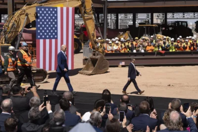 Two individuals walk past an audience and a large U.S. flag at a construction site, with workers in hard hats applauding in the background.