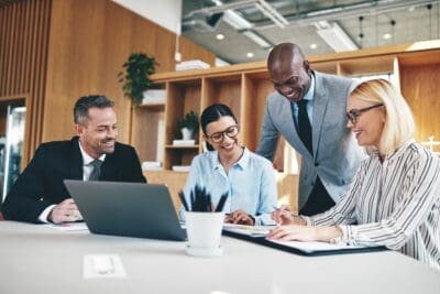 A diverse group of four professionals meeting in an office, sitting around a table with a laptop, papers, and pens. They are engaged and smiling.