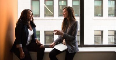 Two women sitting on a windowsill, engaged in conversation, with one holding a laptop, near large windows in an office setting.