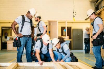 A group of construction workers wearing hard hats and tool belts assemble a wooden frame on the floor in a workshop.