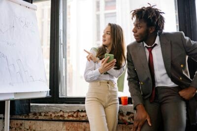Two people in business attire stand by a flipchart with graphs. One points at the chart while holding a phone, and the other leans with a thoughtful expression.
