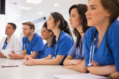 A group of medical professionals in blue scrubs sitting at a table, attentively listening with papers and pens.