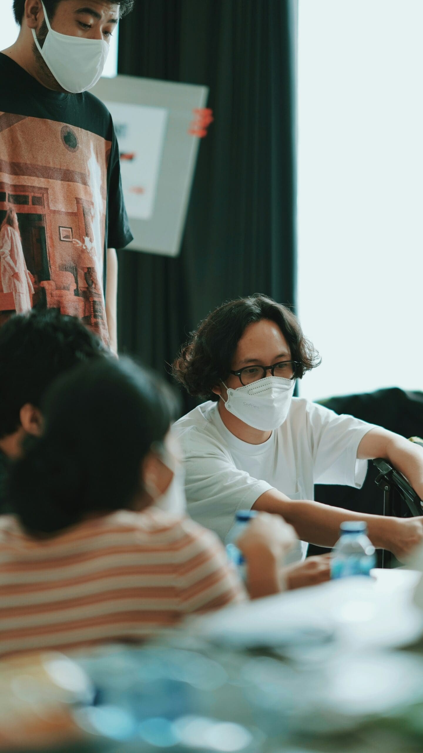 A group of people wearing masks sit and stand around a table with bottled drinks in a well-lit room.