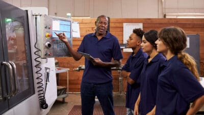 A group of people in blue uniforms listen to a man explaining a machine's control panel in an industrial setting.