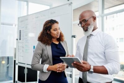 Two professionals in an office discuss content on a tablet, with a whiteboard in the background.