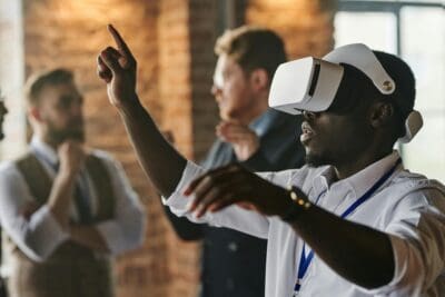 A man wearing a VR headset gestures with his hand, fully immersed in the experience. Three blurred figures linger in the background of the brick-walled room, adding an air of mystery.