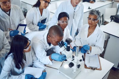 Science, students and medical laboratory with microscope and mentor for pharmaceutical research, education and learning in class. Scientific and chemistry men and women group doing analysis of drug.