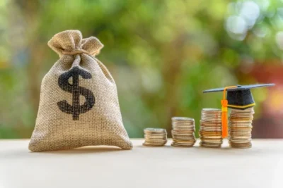 A burlap sack with a dollar symbol next to stacks of coins, one topped with a graduation cap, on a table against a blurred greenery background.