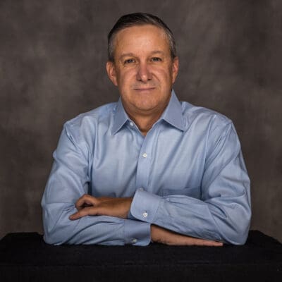 A man in a blue shirt sits with his arms crossed on a black surface, against a dark studio backdrop.