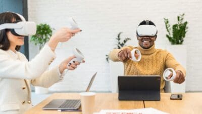 Two people sitting at a table, using virtual reality headsets and controllers, with laptops open. There are plants in the background.