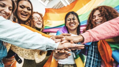 A group of smiling people stand together with hands stacked in front, a rainbow flag in the background.
