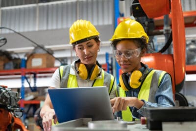 Female automation engineers in safety vest with yellow helmet using laptop for inspection new welding robot arm machine in industrial factory
