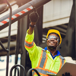 Worker in high-visibility gear and hard hat points upwards, holding a clipboard, with industrial equipment in the background.