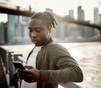 Person wearing earbuds leans on a railing while looking at a smartphone, with a cityscape and bridge in the background.