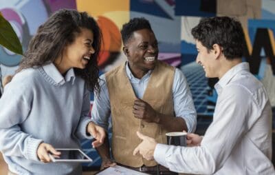 Three young adults engage in a lively discussion, one holding a tablet and another sipping from a mug, in a colorful office setting focused on talent development.