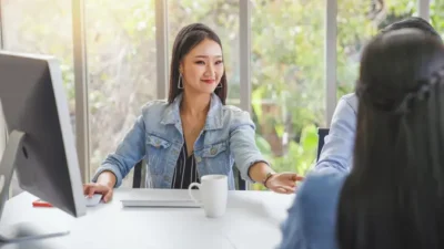 Woman in a denim jacket smiles and extends her hand across a table in an office setting with a computer and a mug, facing another person seated opposite her.