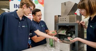 Three people in blue shirts demonstrate a machine converting plastic bottles into green pellets, showcasing recycling technology.