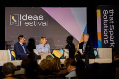 Three people seated on stage in a panel discussion at the Ideas Festival, with a backdrop displaying event branding and audience visible in the foreground.