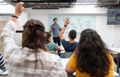 A teacher stands in front of a classroom with a whiteboard. Several students sit facing him, with some raising their hands.