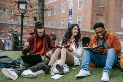Three people sit on grass with books and laptops, smiling and talking against a backdrop of brick buildings.