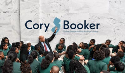 A man in a suit speaks to a group of children seated around him in front of a large sign reading 