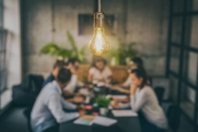 A lit bulb hangs over a blurry conference room where people are having a meeting around a table.