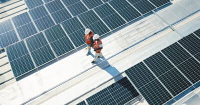 Two workers in safety gear walk between rows of solar panels on a rooftop.