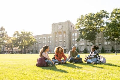 Four people sit in a circle on a grassy field in front of a large building, talking and smiling on a sunny day.