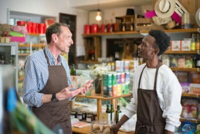 Two men wearing aprons stand in a store, engaged in conversation. Shelves filled with various products are visible in the background.