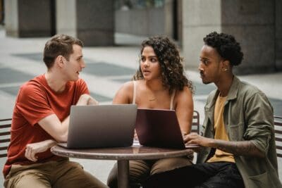 Three people sitting at a table with laptops, engaged in conversation in an indoor setting.