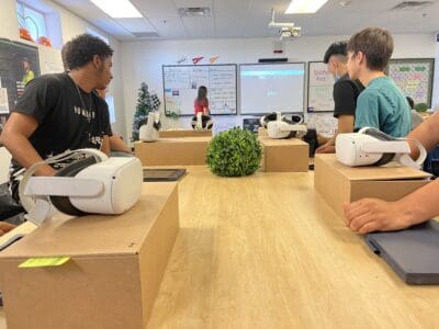 Students sitting around a table with virtual reality headsets and tablets, facing a screen in a classroom.