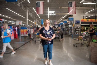 A woman stands smiling in the aisle of a large grocery store, surrounded by shoppers and American flags hanging from the ceiling.