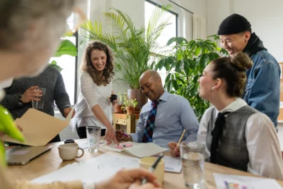 A group of people sit and stand around a table, smiling and interacting, in a room with plants. Some hold papers and a notebook is open on the table.