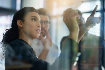 Three future talents are collaborating and brainstorming in front of a glass board, with one person writing or pointing at notes.