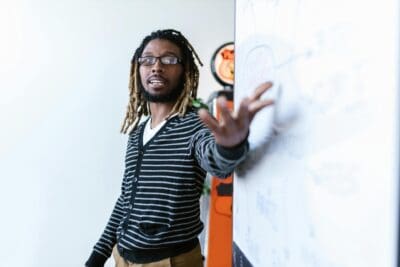 A man in a black-striped cardigan pointing to a whiteboard and presenting on a topic.
