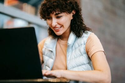 Person with curly hair and a light vest is looking at a laptop screen and smiling. The background is blurred.