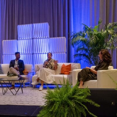 Three people sit on a stage with a blue-lit backdrop and potted plants, engaged in a panel discussion or talk. One person is speaking into a microphone.
