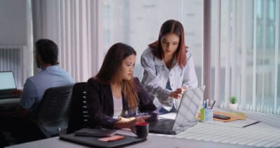 Two women working on a laptop at a desk in a bright office, with a man working on a computer in the background.