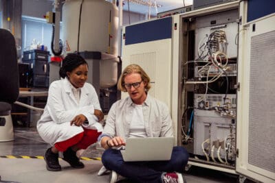 Two scientists in a lab, one sitting with a laptop and the other crouching nearby, discussing in front of open machinery.