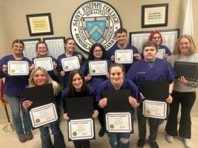 A group of people holding certificates pose in front of a Saint Joseph's College emblem on the wall.