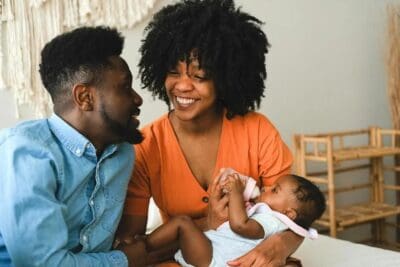 A smiling couple sits together while their baby drinks from a bottle on a couch.