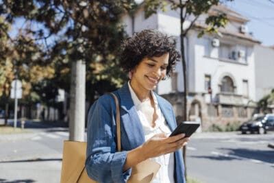 A woman in a blue blazer smiles while looking at her smartphone, standing on a street with trees and buildings in the background.