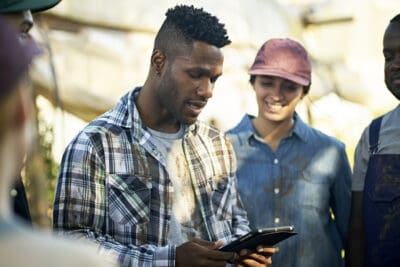 A man in a plaid shirt holds a tablet while speaking to a group of people outdoors. A woman in a cap smiles beside him.